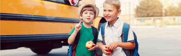 Δύο αστεία χαρούμενα λευκά αγόρια μαθητές παιδιά με μήλα που στέκονται δίπλα στο κίτρινο λεωφορείο την 1η Σεπτεμβρίου ημέρα. Εκπαίδευση πίσω στο σχολείο. Παιδιά έτοιμα να μάθουν και να μελετήσουν. Τίτλος banner ιστού για την ιστοσελίδα. - Φωτογραφία, εικόνα