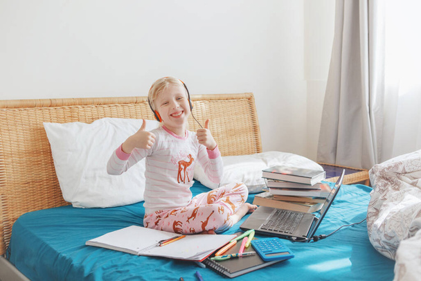 白人の女の子の子供がベッドに座って、ラップトップインターネット上でオンラインで学ぶ。自宅での隔離中にビデオチャット上の仮想クラスレッスン学校。遠隔ビデオ教育クラス.  - 写真・画像