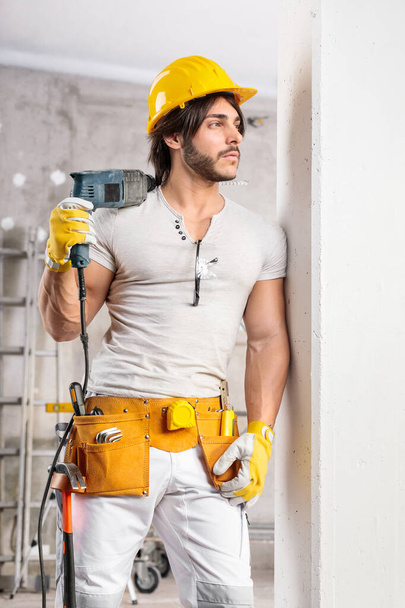 Glücklicher selbstbewusster Bauarbeiter mit Bohrer über der Schulter, der an eine Wand gelehnt in einem Raum im Bau steht und in die Kamera grinst - Foto, Bild