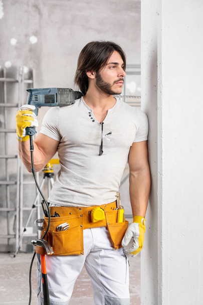Happy confident builder with drill slung over his shoulder standing leaning against a wall in a room under construction grinning at the camera - Φωτογραφία, εικόνα