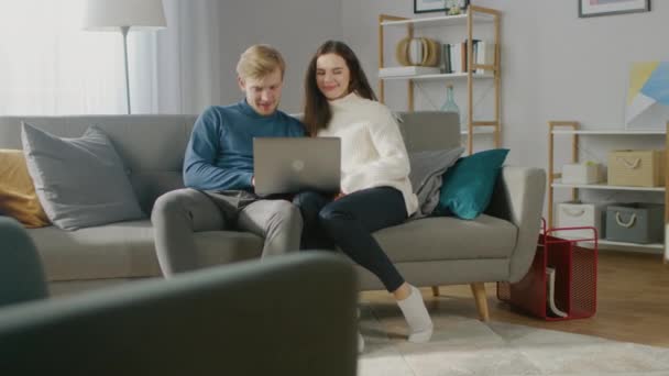 かわいいカップルは、居心地の良いアパートのソファに座っている間、ラップトップコンピュータを使用します。カップルサーフィンウェブ、インターネット上でのショッピング、ソーシャルメディアの使用、ビデオやストリーミングコンテンツの視聴 - 映像、動画