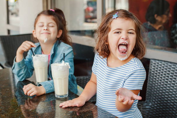 Two funny Caucasian little preschool sisters siblings drink milk shakes in cafe. Friends girls having fun together. Cold summer desserts for kids. Happy authentic childhood lifestyle.  - Fotografie, Obrázek