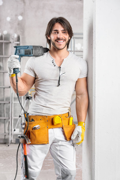 Счастливый уверенный строитель с сверлом, перекинутым через плечо, стоящим, прислонившись к стене в строящейся комнате, ухмыляясь перед камерой
 - Фото, изображение