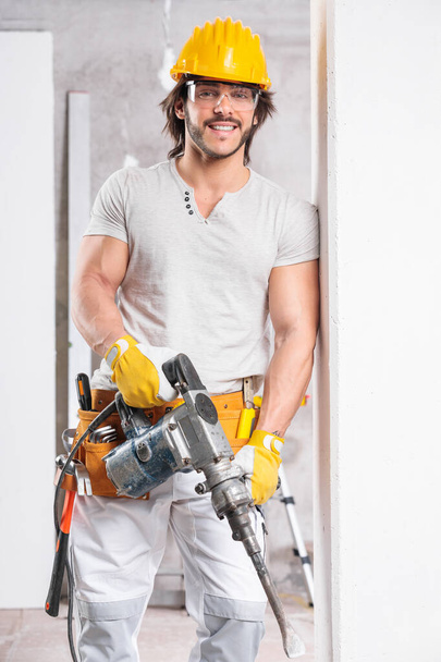 Расслабленный мускулистый строитель в защитном снаряжении и инструментальном поясе, стоящий, опираясь на стену, держа маленький молоток
 - Фото, изображение