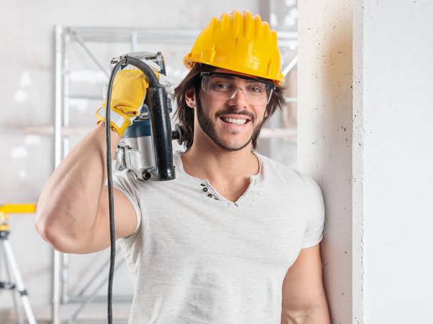 Svalnatý stavitel s bezpečnostními brýlemi a kloboukem, který drží elektrický vrták přes rameno, se opírá o zeď uvnitř a usmívá se na kameru. - Fotografie, Obrázek
