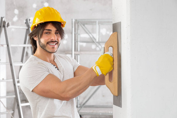 Plasterer finishing the surface of a white wall using a trowel during building or renovations on a house interior and smiling at camera - Photo, Image