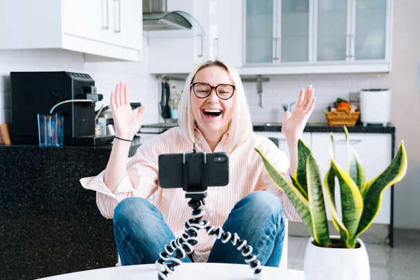 Ευτυχισμένο κορίτσι κάθεται στο σπίτι κουζίνα και κρατώντας videocall. Νεαρή γυναίκα που χρησιμοποιεί smartphone για βιντεοκλήση με φίλο ή οικογένεια. Vlogger καταγραφή webinar. Γυναίκα κοιτάζει κάμερα και χαιρετώντας τα χέρια.  - Φωτογραφία, εικόνα