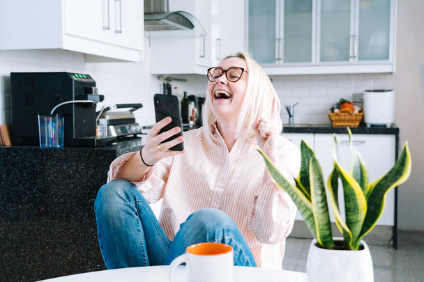Ευτυχισμένο κορίτσι κάθεται στο σπίτι κουζίνα και κρατώντας videocall. Νεαρή γυναίκα που χρησιμοποιεί smartphone για βιντεοκλήση με φίλο ή οικογένεια. Vlogger καταγραφή webinar. Γυναίκα κοιτάζει κάμερα και χαιρετώντας τα χέρια.  - Φωτογραφία, εικόνα