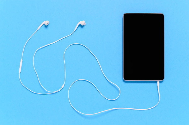 Fekete tabletta fehér fülhallgatóval, kék háttérrel. Minimális koncepció a kapcsolat, távoli digitális munka vagy multimédiás szórakozás, mint a zenei streaming - Fotó, kép