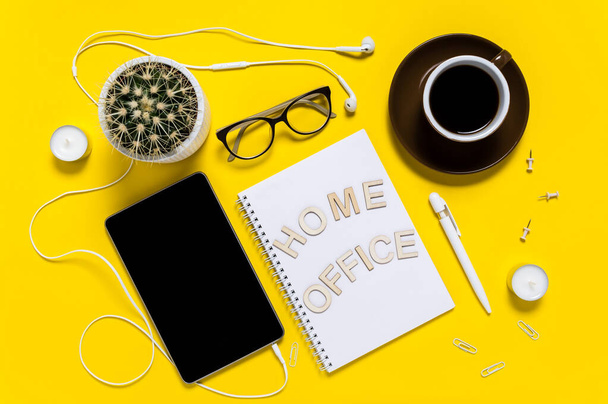 Flat lay of modern workspace with tablet, earphones, coffee cup, notepad, pen, eyeglasses and cactus. Letters saying Home Office. Top view on yellow background - Zdjęcie, obraz