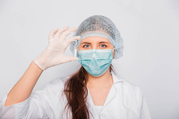 Kadın bir doktorun elinde bir aşı ampulünün yakın çekimi var. Beyaz önlüklü, tıbbi şapkalı, maskeli ve eldivenli laboratuvar teknisyeni elinde ilaç tutuyor. Coronavirus aşısı arama konsepti. - Fotoğraf, Görsel