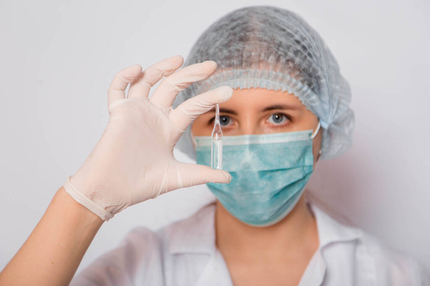 A female doctor in a white coat, medical cap, mask and gloves holds an ampoule with a vaccine. Coronavirus vaccine search concept. - Foto, Imagem