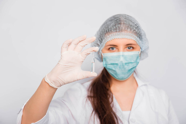 A female doctor in a white coat, medical cap, mask and gloves holds an ampoule with a vaccine. Coronavirus vaccine search concept. - Valokuva, kuva