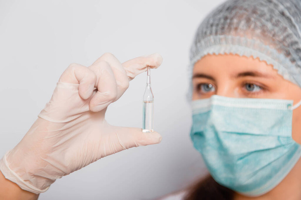 A female doctor in a white coat, medical cap, mask and gloves holds an ampoule with a vaccine. Coronavirus vaccine search concept. - Фото, изображение