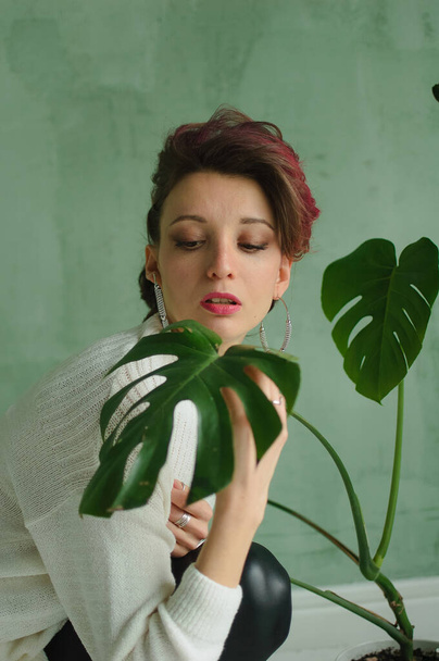 Студійний портрет молодої моделі моди в білому светрі та чорних шкіряних легінсах, що мають в студії з зеленим фоном та екзотичними рослинами
 - Фото, зображення