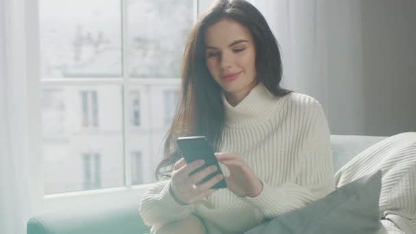 Beautiful Young Woman Using Smartphone Happily, while Sitting on Chair. Sensual Girl Wearing Knitted Sweater, Surfs Internet, Posts On Social Media, Sharing Picture while Relaxing in Cozy Apartment - Materiał filmowy, wideo