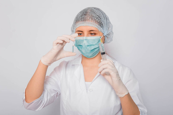 The doctor is holding a syringe and an ampule with a vaccine. Young woman in a white coat, in a medical mask, hat and gloves on a white background. Laboratory medical worker. Coronavirus vaccine search concept. - Foto, afbeelding