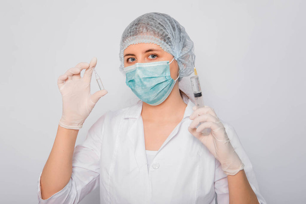 The doctor is holding a syringe and an ampule with a vaccine. Young woman in a white coat, in a medical mask, hat and gloves on a white background. Laboratory medical worker. Coronavirus vaccine search concept. - Valokuva, kuva