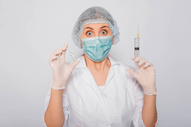 The doctor is holding a syringe and an ampule with a vaccine. Young woman in a white coat, in a medical mask, hat and gloves on a white background. Laboratory medical worker. Coronavirus vaccine search concept. - Fotó, kép