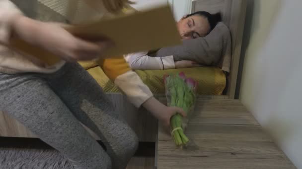 Kindertochter gratuliert überraschter Mutter und legt Blumen und Karte auf Nachttisch - Filmmaterial, Video