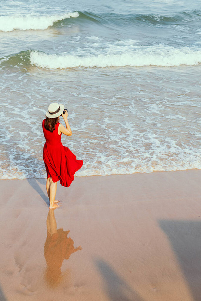 Жінка-фотограф в червоній сукні стоїть і замочує у воді на пляжі і фотографує широку і далеку точку моря. Блюз неба фону і копіювання простору. Ідея способу життя сучасної жінки
. - Фото, зображення