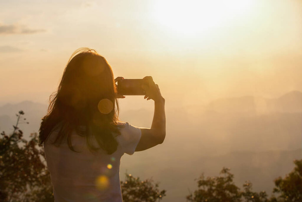 Sunlight flare effects on the back view of female traveler using smartphone mobile to take a photograph of sky and mountains at sunset with orange lights surrounded. Idea for travel in nature place. - Zdjęcie, obraz