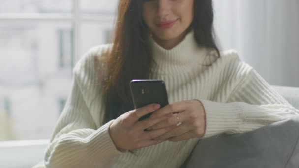 Hermosa mujer joven usando Smartphone sonriendo, mientras se sienta en la silla. Chica sensual con suéter, Navega por Internet, Mensajes en las redes sociales mientras se relaja en acogedor apartamento. Primer plano Retrato
 - Metraje, vídeo
