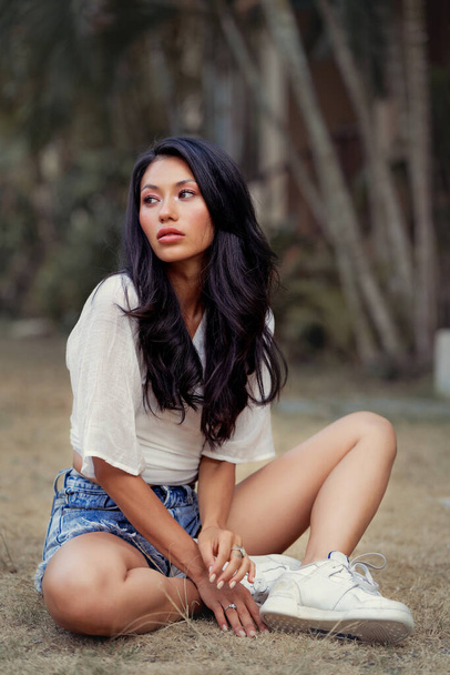Beautiful Asian woman seats on grass, wearing jeans shorts, smiling enjoy weekends - Foto, immagini