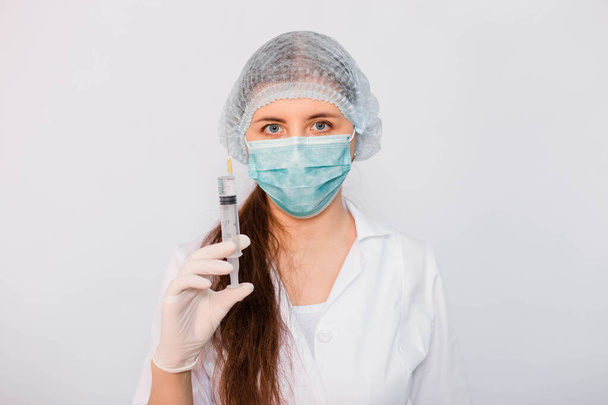 Woman doctor in a white medical coat, mask, hat and gloves holds a syringe with medicine in her hands. Standing on a white background. Coronavirus vaccine search concept. - Fotografie, Obrázek