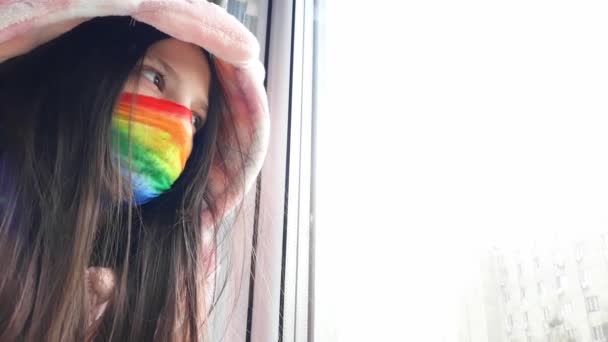 Brunette tiener meisje in een medisch masker geschilderd in heldere regenboog kleuren staat bij het raam met haar hand op glas.Concept van thuis blijven, veilig blijven.Chasetherainbow flash mob.Distance learning. - Video