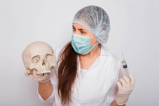 Female doctor in a white medical coat, mask, hat and gloves holds a human skull and a syringe with a vaccine in her hands. The concept of forensic medicine and archeology. Search for a coronavirus vaccine. - Foto, immagini
