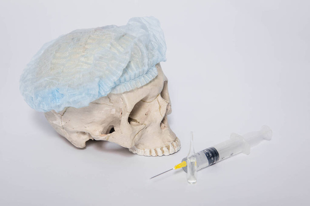 Egy koponya orvosi sapkában egy fecskendő és egy ampulla mellett fekszik, fehér alapon. A törvényszéki orvostudomány és régészet fogalma. Coronavirus vakcina keresése. - Fotó, kép