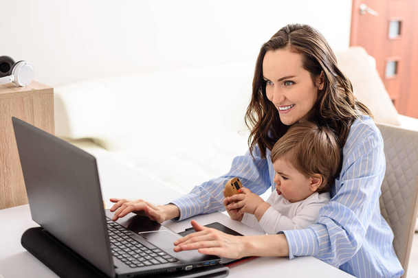 młoda uśmiechnięta brunetka matka w niebieskiej koszuli pracująca na laptopie ze swoim dzieckiem w domu, pracująca na urlopie macierzyńskim - Zdjęcie, obraz