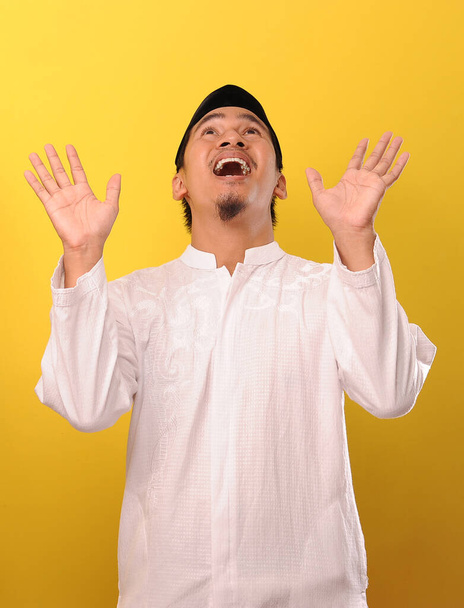 Ein bärtiger junger asiatischer Muslim, der sich mit ausgestreckten Händen davor schützt, dass etwas von oben zu ihm kommt, isoliert auf gelbem Hintergrund - Foto, Bild