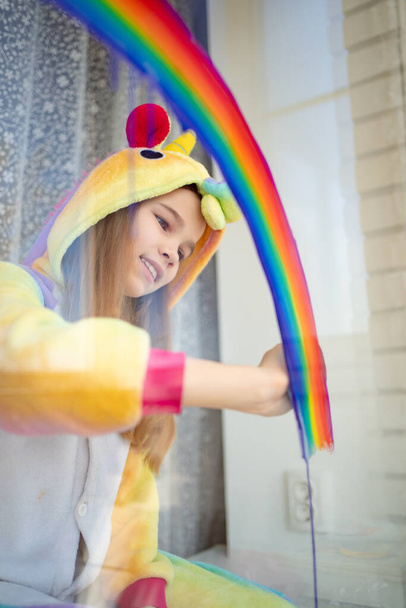 little girl in kigurumi draws rainbow on window. - Zdjęcie, obraz