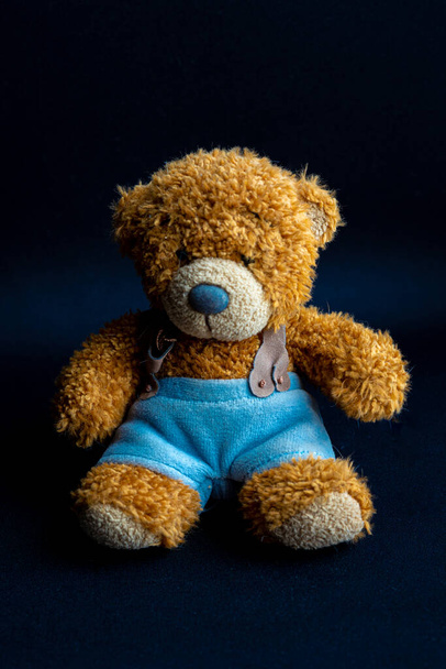 yellow brown teddy bear in blue pants is sitting on dark black background - Foto, Bild