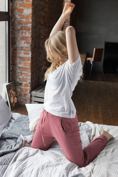 Linda chica vestida con pantalones de pijama rosa, camiseta blanca sentada en la cama con almohada mirando a la ventana con los brazos levantados
. - Foto, Imagen