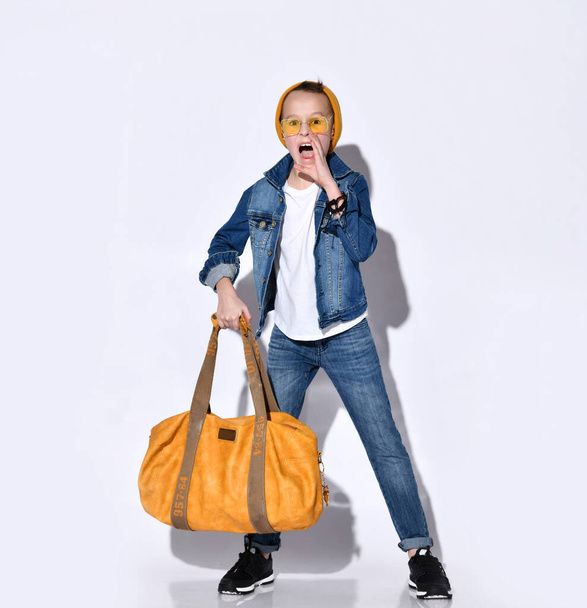 Teenager mit orangefarbener Reisetasche, Hut, Sonnenbrille, T-Shirt, blauem Jeansanzug, schwarzen Turnschuhen. Er posiert isoliert auf Weiß - Foto, Bild