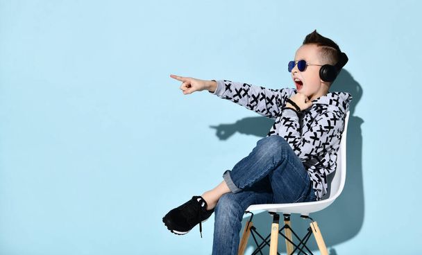 Adolescente em fones de ouvido, óculos de sol e roupas casuais. Apontando para algo, sentado na cadeira branca contra fundo azul
 - Foto, Imagem