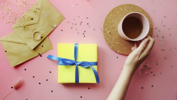 Woman drinking a tea and inspecting a Christmas gift wrapped in yellow paper with blue bow. Top view on wood pink table, flat lay - Footage, Video