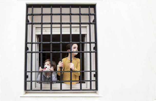 2人の子供は錬鉄製の窓から悲しいことに屋外に見えます。Covid-19監禁の子供への影響 - 写真・画像