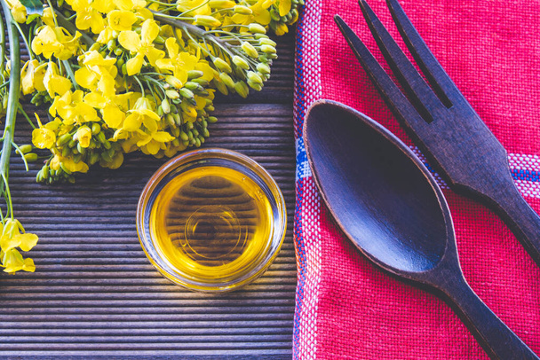 bouquet of spring rape next to a cup with rapeseed oil and wooden cutlery on a wooden table - Фото, изображение
