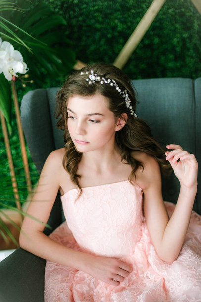 Attractive young girl  in a romantic pink dress by the couch among tropical greenery and white orchids, roses and lilies. Beauty and fashion - Foto, afbeelding