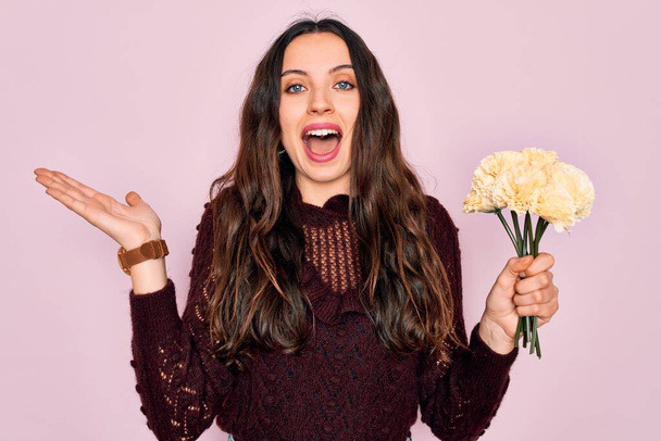 Young beautiful woman with blue eyes holding bouquet of yellow flowers over pink background very happy and excited, winner expression celebrating victory screaming with big smile and raised hands - Photo, Image