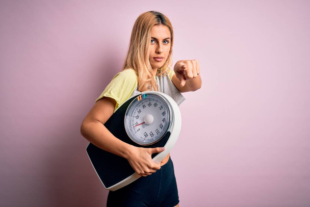 Jonge mooie blonde sportieve vrouw op dieet houden gewicht machine over roze achtergrond wijzend met de vinger naar de camera en naar u, handteken, positieve en zelfverzekerde gebaar van de voorkant - Foto, afbeelding