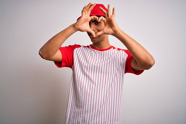 Joven y apuesto deportista afroamericano con camiseta y gorra de béisbol a rayas haciendo forma de corazón con la mano y los dedos sonriendo mirando a través de un cartel
 - Foto, Imagen