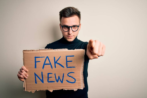 Junger gutaussehender kaukasischer Mann mit Protestkarton für Desinformation und Fake News, der mit dem Finger in die Kamera und auf dich zeigt, Handzeichen, positive und selbstbewusste Geste von vorne - Foto, Bild