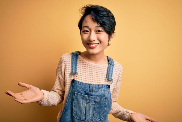 Young beautiful asian girl wearing casual denim overalls over isolated yellow background clueless and confused expression with arms and hands raised. Doubt concept. - Photo, Image