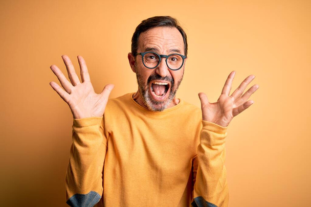 Hombre alzado de mediana edad con suéter casual y gafas sobre fondo amarillo aislado celebrando loco y sorprendido por el éxito con los brazos levantados y los ojos abiertos gritando emocionados. Concepto ganador
 - Foto, Imagen