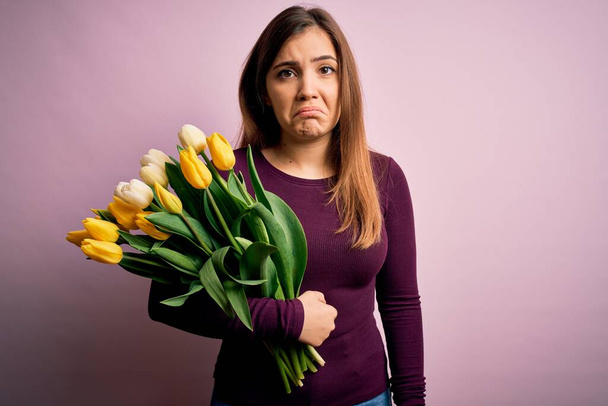 Young blonde woman holding romantic bouquet of yellow tulips flowers over pink background depressed and worry for distress, crying angry and afraid. Sad expression. - Φωτογραφία, εικόνα
